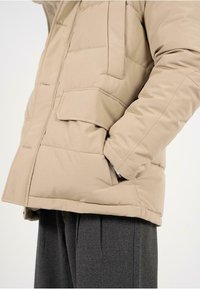 Giacca a vento beige con design trapuntato, dotata di tasca frontale e vestibilità rilassata. Completata da un collo alto e polsini elasticizzati.