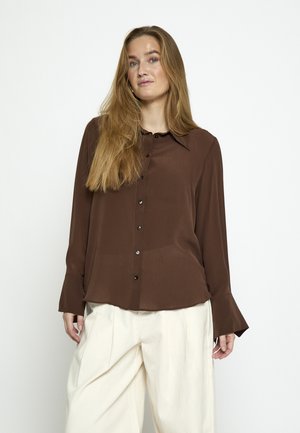 Woman with long blonde hair wearing a brown button-up long-sleeve shirt and loose cream-colored pants against a white background.
