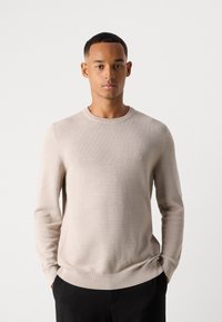 Jeune homme avec les mains dans les poches, portant un pull en maille beige clair et un pantalon noir, sur fond blanc uni.
