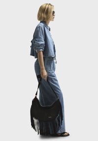 Black suede shoulder bag with long fringe detail, held by a person wearing a cropped denim jacket and wide-leg denim trousers.
