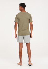 Camiseta de manga corta color verde oliva con un corte relajado, combinada con pantalones cortos gris claro y chanclas, mostrando una vista desde atrás.