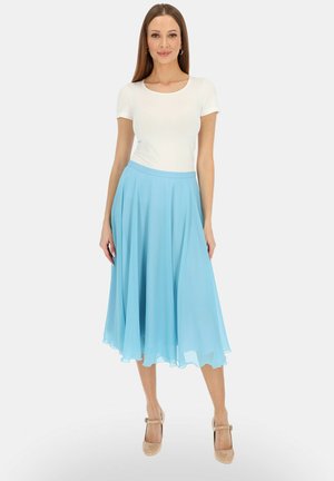 Kobieta ubrana w dopasowany biały krótki rękaw, zwiewną jasnoniebieską spódnicę midi i beżowe szpilki, stojąca na jednolitym tle.