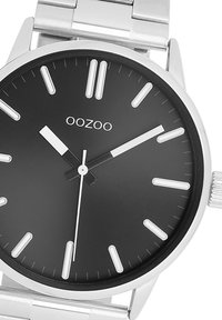Montre en métal argenté ronde avec un cadran noir, des index en argent et des aiguilles blanches distinctes. Dotée d'un bracelet en acier inoxydable. Marque : OOZOO.