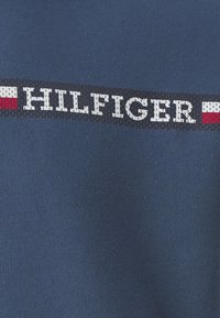 Sudadera de algodón azul con una banda horizontal blanca y roja con el logo que presenta "HILFIGER" en una tipografía con negrita. Textura suave, ajuste informal.