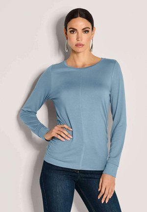 Blaues Langarmoberteil mit rundem Ausschnitt, weichem Stoff, seitlichen Raffungselementen und engschaftigen Ärmeln. Kombiniert mit dunklen Jeans.
