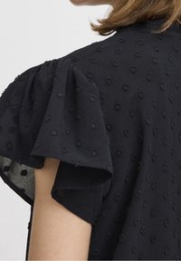Schwarze Bluse mit Textur, die Puffärmel und einen weichen Stoff mit geprägten Punktmustern aufweist, von hinten betrachtet.