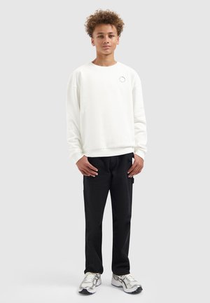 Tienerjongen met krullend haar staat, draagt een wit longsleeve shirt, zwarte broek en witte sneakers op een effen lichte achtergrond.