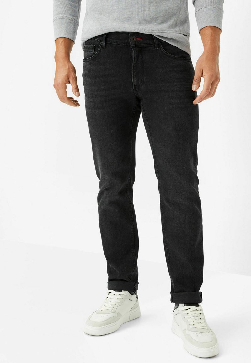 Schwarze Denim-Jeans mit Slim-Fit, ausgestattet mit fünf Taschen und einem dezenten roten Logoakzent. Getragen mit weißen Sneakers, hochgerollten Säumen sichtbar.