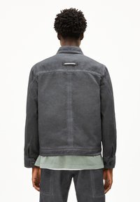 Graue Denimjacke mit lockerem Schnitt, sichtbaren Nähten, Knopfmanschetten und einem Logostick auf der Rückseite. Darunter ein grünes Hemd.