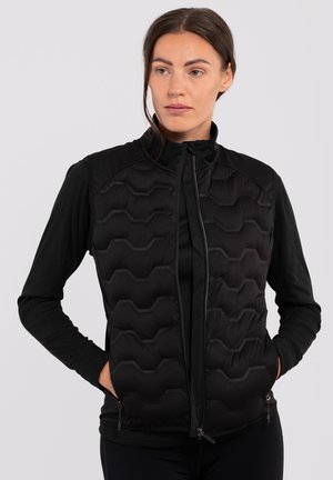 Black vest with hexagonal quilted pattern and zip closure, featuring textured shoulders and side pockets. Worn over a long-sleeve top.