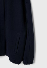 Détail de la manche et de la poche d'une veste en laine bleu marine foncé avec un poignet élastique sur un fond gris clair.