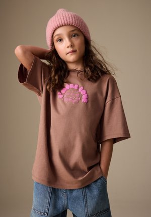 Jong meisje draagt een roze gebreide muts, een bruin T-shirt met de tekst "S'more Love" en een blauwe spijkerbroek, poserend met een hand achter haar hoofd.