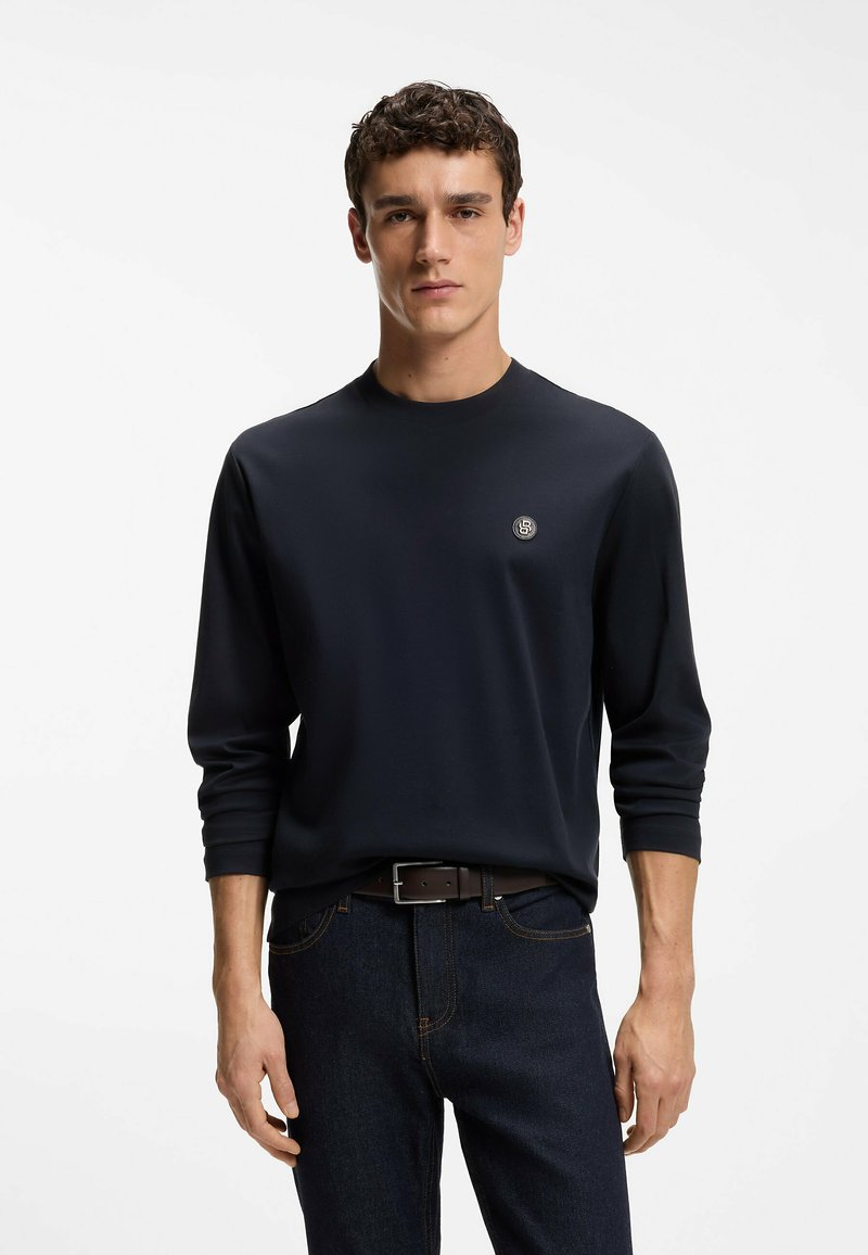 Jeune homme portant une chemise bleu marine à manches longues avec un petit logo rond sur la poitrine et un jean bleu foncé avec une ceinture noire.