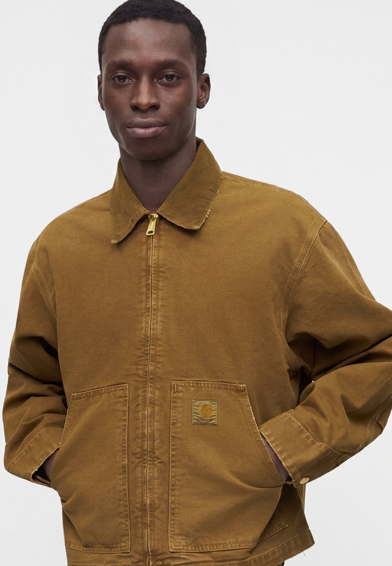 Young man with dark skin wearing a brown zip-up jacket with front pockets and standing with hands in pockets.
