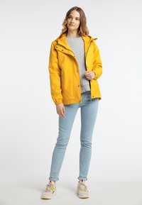 Gul vattentät jacka med huva, tryckknappar, elastiska ärmslut och en avslappnad passform, i kombination med ljusblåjeans och neutrala sneakers.