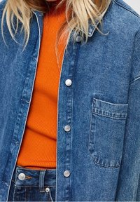 Blå denimjacka med silvriga tryckknappar och ett bröstfick, lager på lager över en orange ribbad tröja. Har en avslappnad passform och textilstruktur.