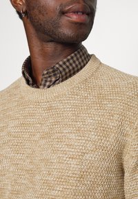 Maglione beige a trama con scollo rotondo, indossato sopra una camicia a quadri marrone scuro. Il pattern a maglia aggiunge interesse visivo e profondità.