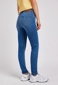 Personne portant un jean bleu ajusté Lee, un haut jaune et des baskets blanches, debout devant un fond clair uni.