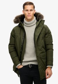 Parka acolchada de color verde oliva con capucha forrada de piel sintética, cierres de cremallera y a presión, bolsillos laterales, llevada sobre un suéter de punto de canalé gris claro con cuello alto.