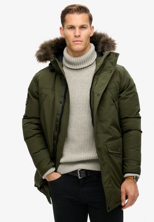 Olive green padded parka jacket with a faux fur-lined hood, zip and snap closures, side pockets, worn over a light grey ribbed turtleneck sweater.