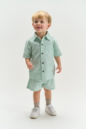 Fiatal gyermek áll, zöld-fehér csíkos rövid ujjú pólót és hozzáillő rövidnadrágot visel fehér tornacipővel és bézs zoknival.