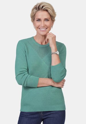 Glimlachende vrouw met kort blond haar, een groene textuur trui en horloge dragend, poseren met haar hand dichtbij de kin tegen een witte achtergrond.