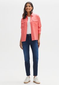 Korallenfarbenes Button-Down-Hemd mit zwei Brusttaschen, kombiniert mit dunkelblauen Jeans und weißen Sneakern. Glatte Stoffqualität mit lockerem Schnitt.