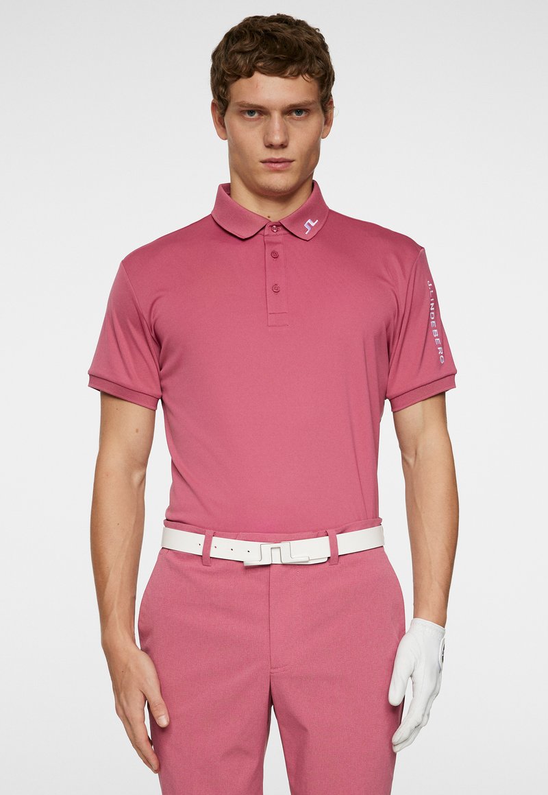 Rosa Poloshirt mit Kragen, kurzen Ärmeln und subtiler Textur. Ausgestattet mit einem weißen Gürtel und einem Golfhandschuh; Logo auf dem Ärmel sichtbar.