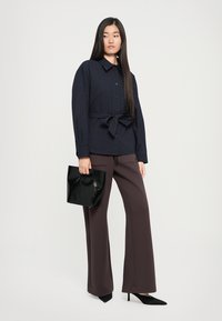 Navy striped shirt with button front and tie waist, paired with wide-leg brown trousers. Model holds a shiny black clutch purse.