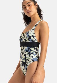 Maillot de bain une pièce à motif floral multicolore, avec une bande noire à la taille, des bretelles larges et des jambes hautes. Texture de tissu lisse.