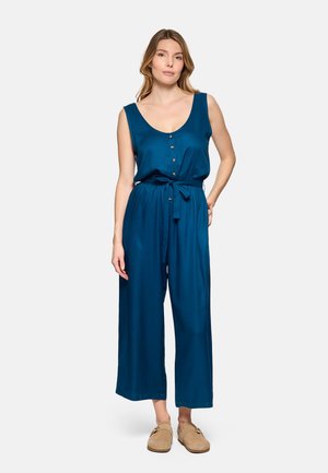 Mujer de pie con mono azul sin mangas de botones al frente y lazo en la cintura, y zapatos beige sin cordones, sobre fondo claro.