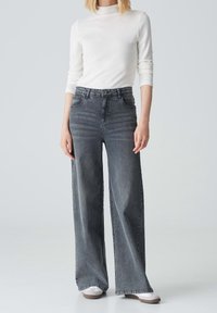 Mujer con una camiseta blanca de manga larga, jeans anchos de tiro alto en negro desgastado y zapatillas blancas, de pie frente a un fondo simple.