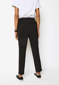 Schwarze tapering Hosen mit elastischem Bund, ausgestattet mit zwei Gesäßtaschen und einer glatten Stoffoberfläche. Kombiniert mit schwarzen Sneakers.