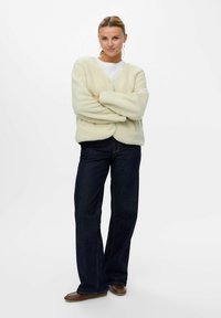 Cardigan crème, fluffy, avec des manches longues et des boutons à l'avant, associé à un jean flare bleu foncé et des ballerines marron.