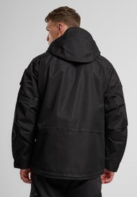 Giacca nera impermeabile con cappuccio, caratterizzata da una vestibilità ampia, tasche laterali e polsini regolabili. Texture liscia e design minimalista.