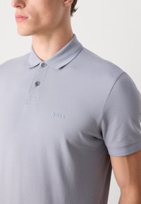 Camiseta polo gris con una textura suave, con tapeta de tres botones y un sutil bordado del logo en el pecho. Las mangas cortas están terminadas de manera ordenada.