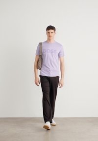 T-shirt lavande avec l'inscription "BOSS", pantalon noir et chaussures blanches. Le modèle porte un sac à épaule marron clair et se tient sur un sol neutre.
