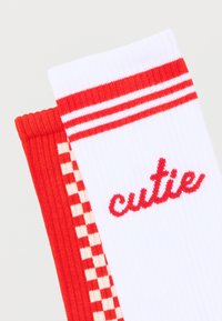 Δύο διπλωμένες κάλτσες, η μία λευκή με κόκκινες ρίγες και το κείμενο "cutie", η άλλη κόκκινη με λευκό μοτίβο καρό.