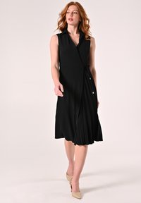 Zwarte mouwloze jurk met een overslagontwerp, met plooien in de rok, een kraagachtige halslijn en een ceintuurelement in de taille. Glad stofafwerking.