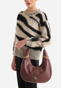 Borsa hobo in pelle marrone dalla texture liscia con accenti dorati, tenuta da una persona che indossa un maglione a righe nere e beige.