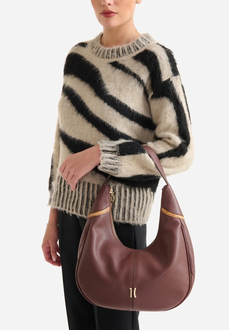 Borsa hobo in pelle marrone dalla texture liscia con accenti dorati, tenuta da una persona che indossa un maglione a righe nere e beige.
