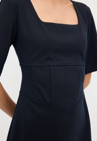 Abito blu navy con scollatura quadrata, maniche corte e corpino strutturato. Presenta cuciture angolari e un tessuto liscio e strutturato.