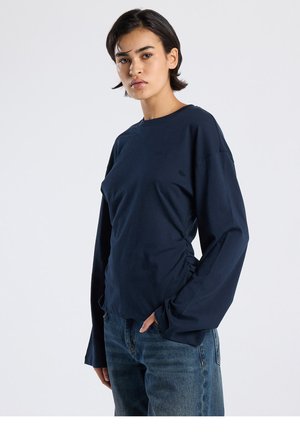 Tummansininen pitkähihaispaita, valmistettu pehmeästä puuvillasta, vartalonmyötäinen malli sivutaitoksilla. Yhdistetty rentoihin farkkuihin.