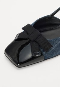 Chaussure plate avec un corps en denim foncé, un embout en vernis noir et un détail de nœud en ruban noir. Présente une forme de bout carré et un design à enfiler.