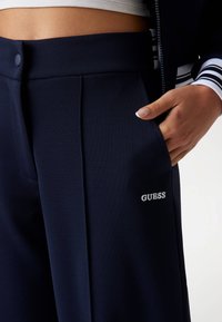 Marineblauwe broek met een op maat gemaakt ontwerp, voorzien van een knoopsluiting en zichtbare zak. Het "GUESS"-logo is in het wit geborduurd op de voorkant.