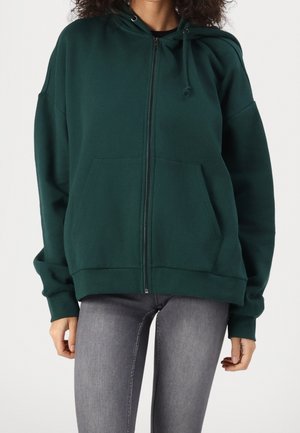 Femme portant un sweat à capuche vert foncé zippé avec poches avant et un jean skinny gris sur un fond uni.