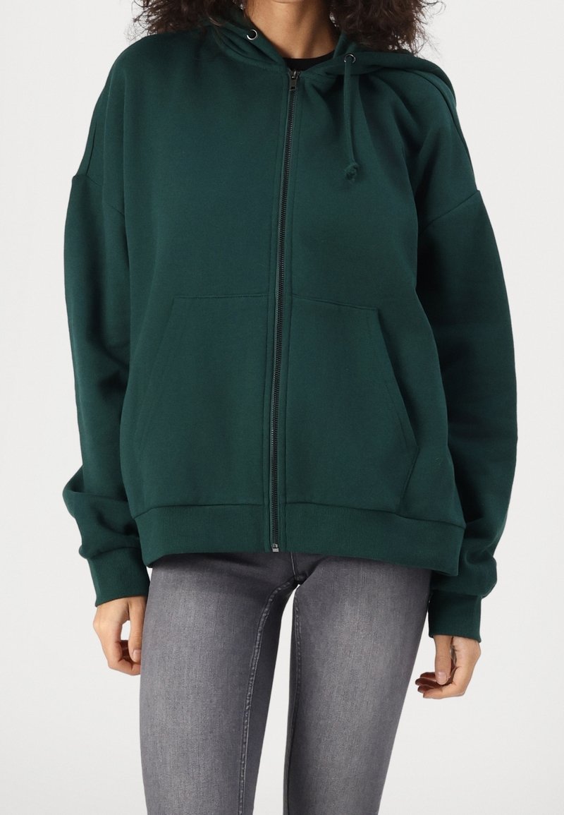 Femme portant un sweat à capuche vert foncé zippé avec poches avant et un jean skinny gris sur un fond uni.