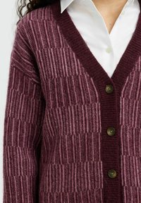 Teksturowany burgundowy kardigan z dekoltem w serek, pionowym wzorem prążków oraz trzema brązowymi guzikami z przodu. Połączony z białą koszulą pod spodem.