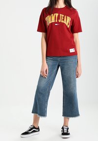Camiseta de algodón roja con el logo "TOMMY JEANS" en amarillo, combinada con unos vaqueros de pierna ancha azul. Zapatillas negras con cordones blancos.