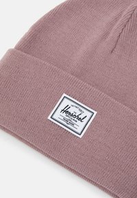 Berretto lavorato a maglia rosa realizzato in morbido filato, con un'etichetta rettangolare bianca con testo nero, che indica i dettagli del marchio e del marchio registrato.